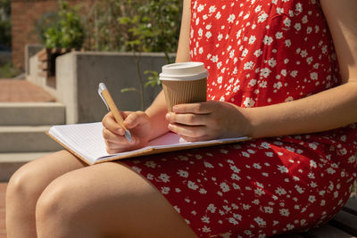 Unrecognizable young woman in red dress drinking coffee from craft paper cup writing gratitude