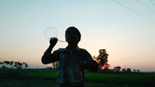 Boy with bucket blowing bubble on field during sunset