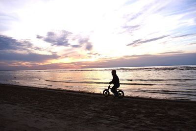 Silhouette boy riding bicycle at beach during sunset