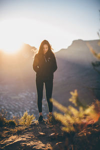 Young woman with hands in pockets standing on mountain against sky during sunset