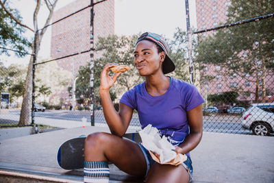 Smiling young woman eating food while sitting on street on road