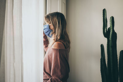 Young woman looking outside the window with surgical mask during coronavirus lockdown virus covid-19