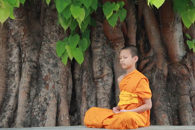 Monk praying while sitting by tree trunk