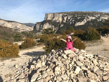 Happy girl with arms raised sitting on rocks against sky during sunny day