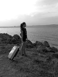 Side view of young woman with luggage standing at beach against sky during sunset