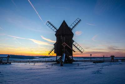 Traditional windmill on snow covered land against sky during sunset