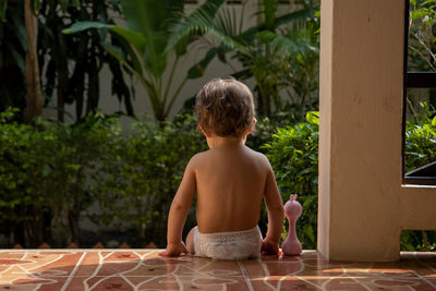 Rear view of shirtless girl sitting against plants