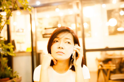 Close-up of thoughtful young woman talking on phone in illuminated cafe