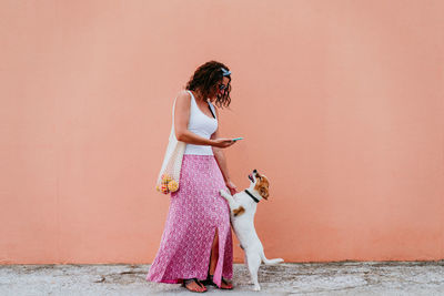 Full length of woman with dog holding phone while standing on footpath against wall