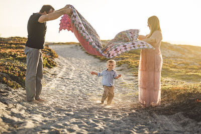 Side view of parents holding blanket over cute son running on sand at beach against clear sky during sunset