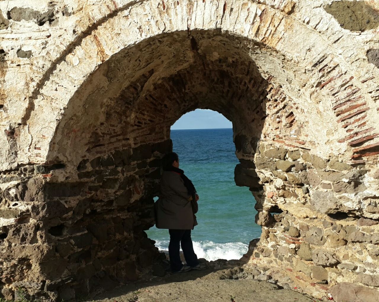 arch, real people, rock - object, rock formation, standing, one person, architecture, nature, sea, full length, lifestyles, day, leisure activity, built structure, beauty in nature, rear view, women, natural arch, men, water, scenics, sky, outdoors, people