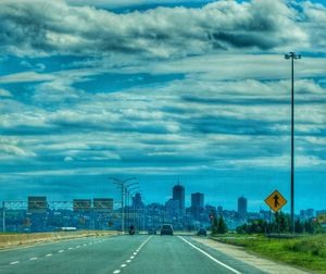 Road passing through city against cloudy sky