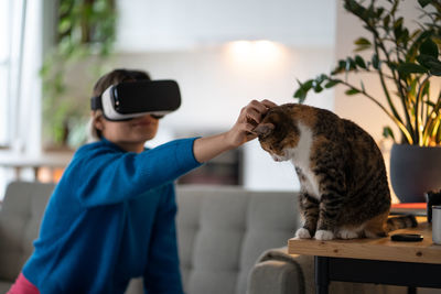 Positive woman in vr helmet petting cat sits on couch watching movie with virtual reality effects