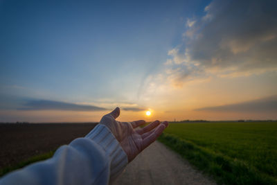 Close-up of person hand over field against sky during sunset