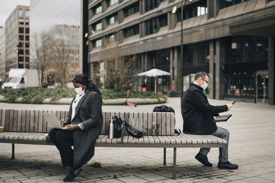 Businessman with smart phone while female colleague using laptop sitting on bench during covid-19