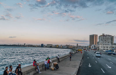 People on road by buildings against sky during sunset