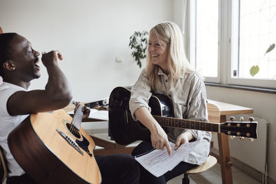 Happy female tutor looking at male student laughing while practicing guitar in classroom