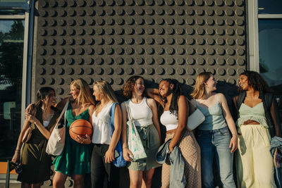 Happy teenage girls looking at each other while standing against wall during sunny day