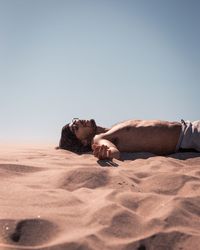 Shirtless man lying on sand against clear sky