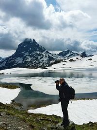 Hiker photographing while standing at lakeshore against snowcapped mountains