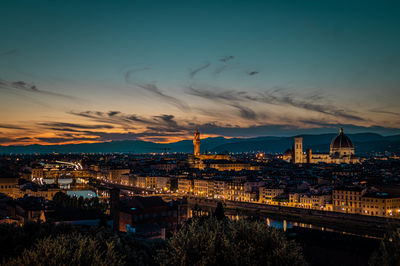 Illuminated bridge over river by buildings against sky during sunset