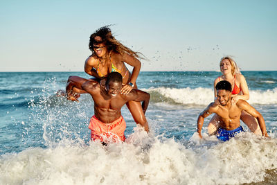 Happy multiracial women climbing on backs of black boyfriends while playing in foamy sea waves on summer day