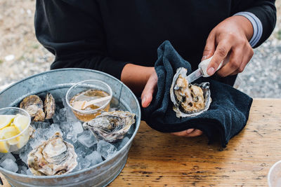 Woman shucking oyster at table outdoors with ice-filled tub of oysters and condiments