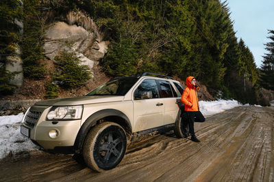 Side view of man in orange jacket standing by off-road car on muddy road in winter