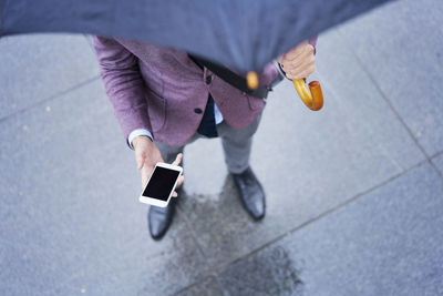 Businessman standing under umbrella holding cell phone, partial view