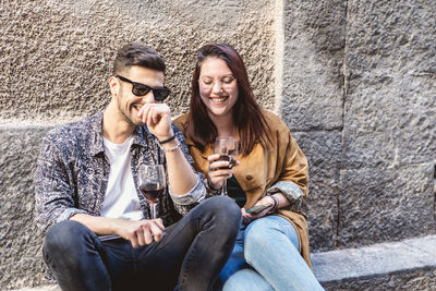 Young woman and man holding drink in glass while sitting against wall