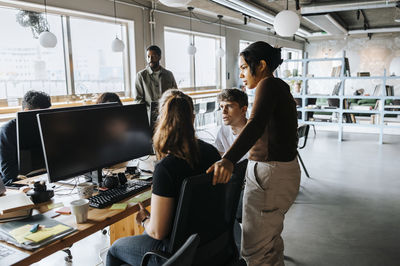 Young female entrepreneur standing by colleagues discussing while sitting on chair at office