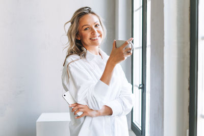 Young smiling blonde woman with long hair in white shirt rest and drinking tea near window in office
