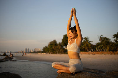 Portrait of young woman sitting at beach against sky during sunset