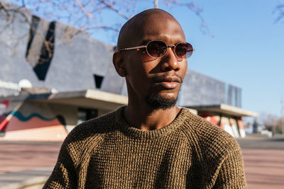 Confident african american male in fashionable sunglasses and sweater sitting on street with leafless trees on sunny day in city