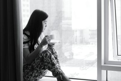 Woman drinking coffee while looking through window at home