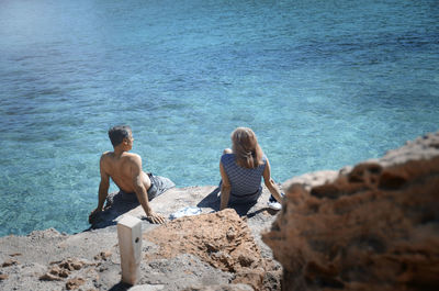 Rear view of couple sitting on rock by sea