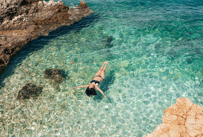 High angle view of young woman in black bikini swimming in transparent sea water.