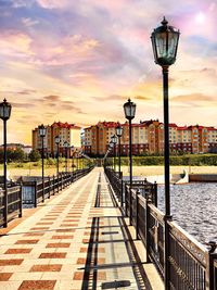 Street lights on footbridge over river in city against sky during sunset