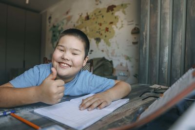 Smiling asian boy show thumb up in home study room with world map, education blog, learning platform