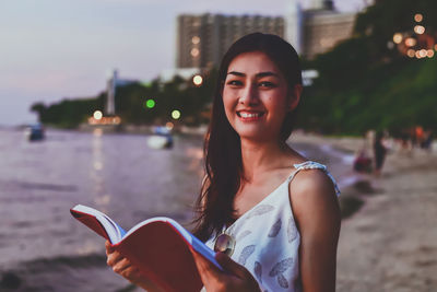 Portrait of smiling young woman holding book at beach in city