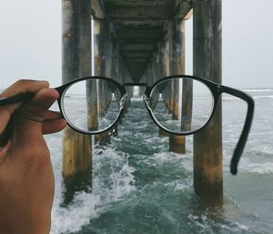 Cropped hand holding eyeglasses under bridge on sea