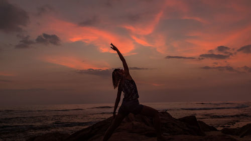 Rear view of woman stretching at rocky shore against orange sky