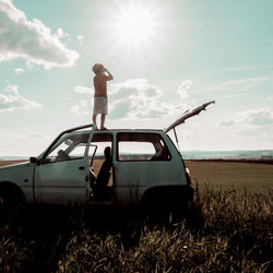 Low angle view of carefree boy standing on car roof at field against cloudy sky during sunny day