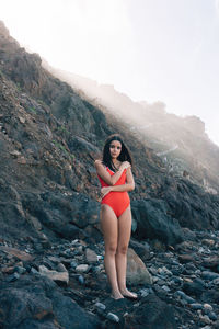 Full body of slim female in red swimwear looking at camera while standing on stony shore near rock in coastal area