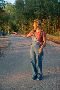 Brazilian millennial girl in denim overalls stands on forest road,points with her hand at sunset