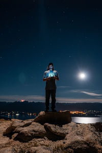Full length of man operating drone while standing on rock against sky at night