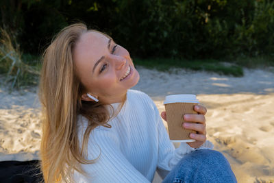 Happy young woman drinking coffee from paper cup takeaway on the beach sea ocean. wearing wireless