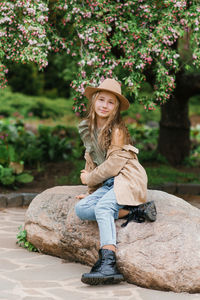 Cute teenage girl with long brown hair in a hat and raincoat is sitting on a rock in the park