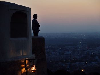 Man looking at illuminated cityscape against sky during sunset