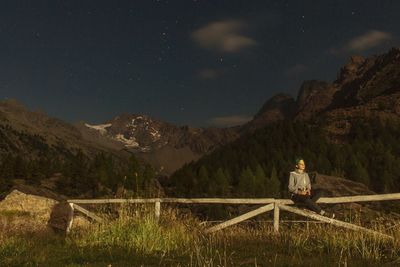 Man sitting on fence against mountain at night